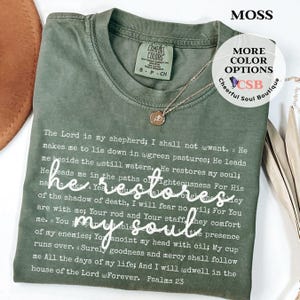 Puede incluir: Una camiseta de cuello redondo color verde musgo con el texto completo del Salmo 23 impreso en blanco, con la frase 'he restores my soul' en una fuente cursiva blanca grande en el centro. La camiseta tiene una etiqueta de Comfort Colors en el cuello.