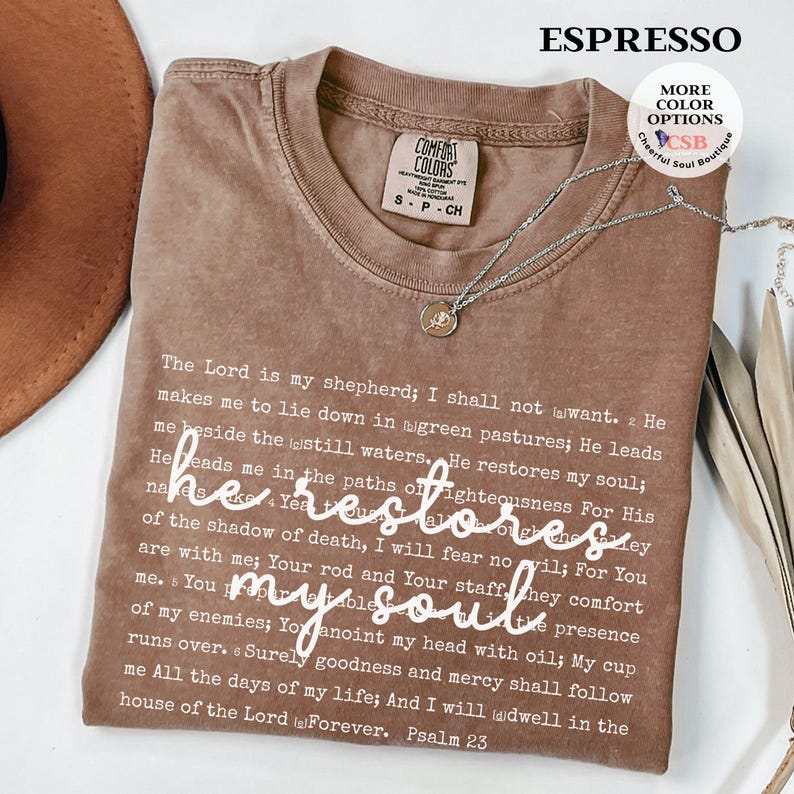 Puede incluir: Una camiseta de cuello redondo color marr&oacute;n con el texto del Salmo 23 impreso en fuente blanca peque&ntilde;a, y la frase he restores my soul escrita en una fuente cursiva blanca m&aacute;s grande en el centro. Un collar plateado descansa sobre la camiseta.