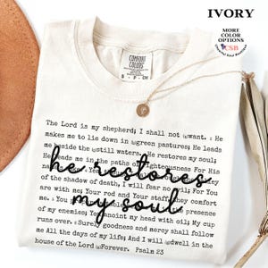Puede incluir: Una camiseta color marfil con el texto completo del Salmo 23 impreso en fuente negra estilo m&aacute;quina de escribir. La frase 'he restores my soul' est&aacute; superpuesta en una letra cursiva negra grande. Un collar dorado con un peque&ntilde;o dije redondo descansa encima.