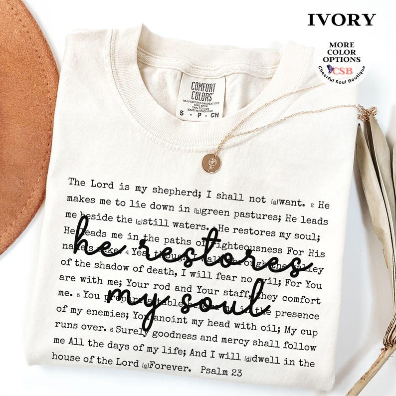Puede incluir: Una camiseta color marfil con el texto completo del Salmo 23 impreso en fuente negra estilo m&aacute;quina de escribir. La frase 'he restores my soul' est&aacute; superpuesta en una letra cursiva negra grande. Un collar dorado con un peque&ntilde;o dije redondo descansa encima.