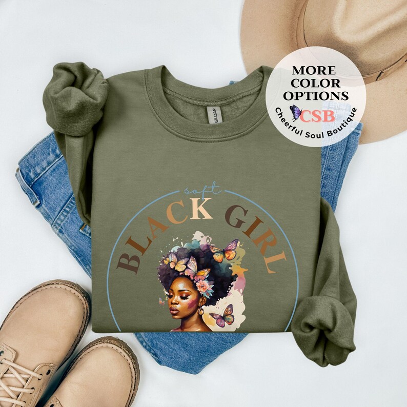 May include: A green sweatshirt with a black circle graphic that says "soft black girl" in a stylized font. The graphic also features a black woman with brown hair and butterflies.