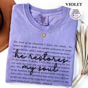 Puede incluir: Una camiseta violeta de manga corta con el texto completo del Salmo 23 impreso en letra negra peque&ntilde;a, con la frase 'he restores my soul' escrita en una fuente cursiva negra m&aacute;s grande en el centro. La camiseta tiene un cuello redondo y etiqueta.