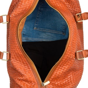 May include: A brown leather woven duffel bag with a gold zipper open to reveal a pair of blue jeans and a black garment inside.