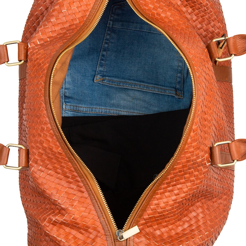 May include: A brown leather woven duffel bag with a gold zipper open to reveal a pair of blue jeans and a black garment inside.