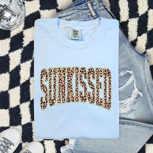 以下が含まれることがあります： ブラウンとブラックのヒョウ柄で、太字のアーチ状フォントで「SUNKISSED」という文字がプリントされたライトブルーのクルーネックTシャツ。首元の内側にはComfort ColorsブランドのLサイズを示すタグが付いています。