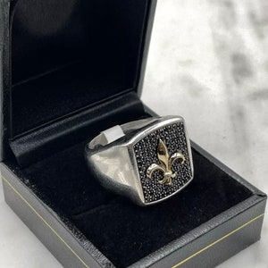 May include: A silver ring with a black stone and a gold fleur-de-lis design. The ring is in a black velvet box.
