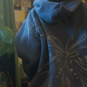 May include: A blue hoodie with a white floral design on the back. The hoodie has a hood and long sleeves. The design is a stylized floral pattern with star-like accents. The hoodie is made of a soft material.