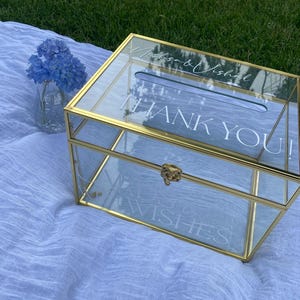 May include: A clear glass and gold metal wedding card box with the words "Thank You" and "Wishes" printed on the glass. The box has a slot on top for cards and a decorative clasp. A vase of blue flowers sits nearby.