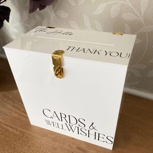 May include: A white acrylic card box with gold-colored hardware. The box has a slot on top and features the words "THANK YOU!" and "CARDS & WELL WISHES" in black script. The top of the box is inscribed with "The Halls".