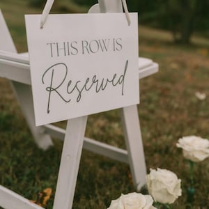 May include: White sign hanging on a white chair with the words "THIS ROW IS Reserved" in elegant script. The sign is at a wedding or event, with white roses in the foreground.