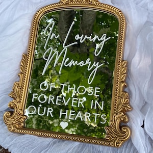 May include: A gold-framed mirror with ornate detailing. The mirror reflects a green, leafy background. White text reads "In Loving Memory of Those Forever in Our Hearts" with a heart symbol.