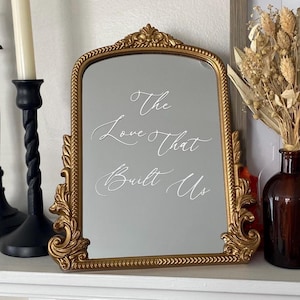 May include: A gold-framed mirror with the inscription "The Love That Built Us" in elegant white script. The mirror has an ornate, vintage design and is displayed on a white surface. Decorative elements include a black candlestick and a vase with dried flowers.