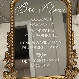 May include: A gold-framed mirror with a decorative, ornate design. The mirror displays a "Bar Menu" in elegant script, listing cocktail options like Coconut, Margarita, and Blueberry Smash. The overall aesthetic is vintage and luxurious.