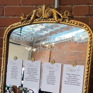 May include: A gold-framed mirror with the text "WE'RE SO glad YOU'RE HERE, your seat awaits" etched on the glass. Four white cards with guest names are attached. The mirror has an ornate, decorative top.