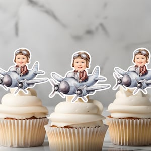 Op de afbeelding: Drie cupcake-toppers met een cartoonillustratie van een kind dat verkleed is als piloot in een vliegtuig. De toppers zijn gedrukt op wit karton en zijn klaar om op cupcakes te worden geplaatst.