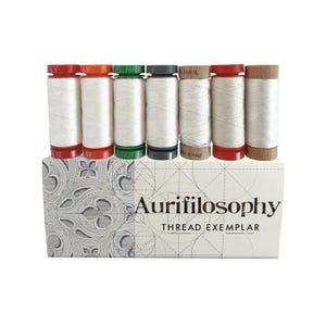 May include: A collection of white thread spools with various colored caps, including red, orange, green, and brown, are displayed on top of a decorative box. The box features the text "Aurifilosophy THREAD EXEMPLAR" and a patterned design.