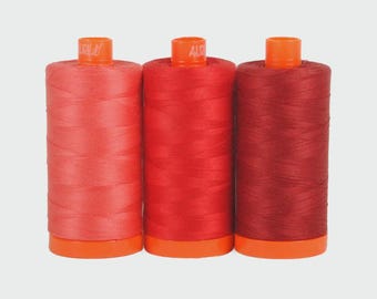 May include: Three spools of thread in shades of red, from light coral to deep burgundy. Each spool is cylindrical, with an orange base and top. The thread is tightly wound, and the spools are arranged side by side against a neutral background.