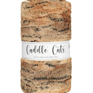 May include: A rolled-up bundle of brown and tan faux fur fabric. A white band around the middle reads "Cuddle Cuts" and "2 Yard Cuddle minky fabric precut". The fabric has a soft, textured appearance, suitable for various sewing projects.