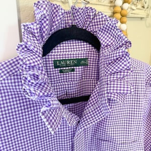 May include: A purple and white gingham button-down shirt featuring a prominent ruffled collar. The interior label reads LAUREN RALPH LAUREN, CLASSIC FIT, NON IRON, with size 16 1/2 34/35. The shirt is displayed on a black hanger.