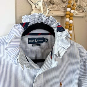 May include: A light blue and white striped button-down shirt featuring a ruffled collar with navy and red striped trim. The interior label reads Ralph Lauren Classic Fit size M. A small embroidered logo is visible on the lower front of the shirt.