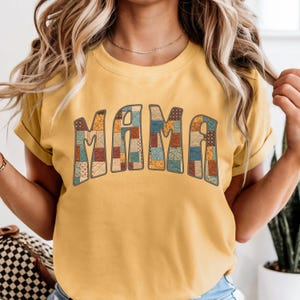May include: Mustard yellow t-shirt with the word "MAMA" in a patchwork quilt design. The letters are filled with various patterns in shades of brown, blue, and orange. The shirt has rolled-up sleeves.