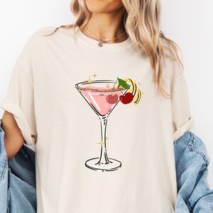 May include: Cream-colored t-shirt featuring a pink cocktail in a martini glass. The cocktail is garnished with cherries and a lemon twist. The design includes gold star accents, creating a fun and stylish graphic tee.