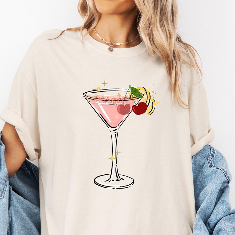 May include: Cream-colored t-shirt featuring a pink cocktail in a martini glass. The cocktail is garnished with cherries and a lemon twist. The design includes gold star accents, creating a fun and stylish graphic tee.