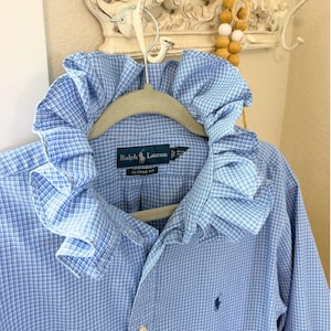 May include: A blue and white checkered button-down shirt featuring a large, ruffled collar. The shirt has a Ralph Lauren label on the inside neck area with the text Classic Fit, and a small navy blue embroidered logo on the lower front section.