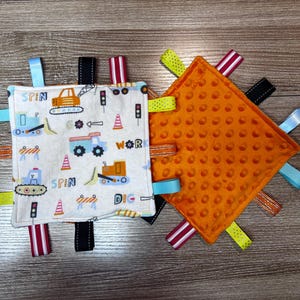 May include: A white and orange baby sensory toy with a variety of textures and colors. The toy features a variety of ribbons and tags, as well as the words "Spin", "Work", "Dig", and "Spin".