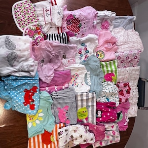 May include: A collection of baby clothes in various colors and patterns, including pink, blue, and white. Several shirts feature graphic designs, such as strawberries, angels wings, and the text "Jesus Loves Me".