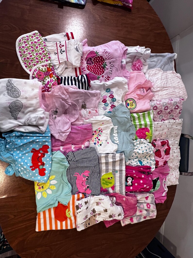 May include: A collection of baby clothes in various colors and patterns, including pink, blue, and white. Several shirts feature graphic designs, such as strawberries, angels wings, and the text "Jesus Loves Me".