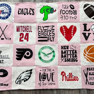 May include: A pink and white patchwork quilt featuring various sports-themed designs. Includes logos for the 76ers, Eagles, Phillies, and Flyers, along with phrases like "Eat Sleep Football Repeat" and "For the Love of the Game."