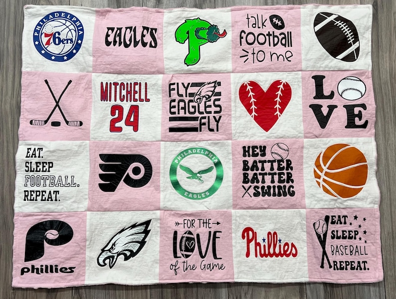 May include: A pink and white patchwork quilt featuring various sports-themed designs. Includes logos for the 76ers, Eagles, Phillies, and Flyers, along with phrases like "Eat Sleep Football Repeat" and "For the Love of the Game."