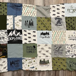 May include: A patchwork quilt made from various t-shirts, featuring camping-themed graphics and text. The quilt includes images of tents, trees, bears, and phrases like "Explore" and "Happy Camper". The color palette includes greens, blues, and grays.