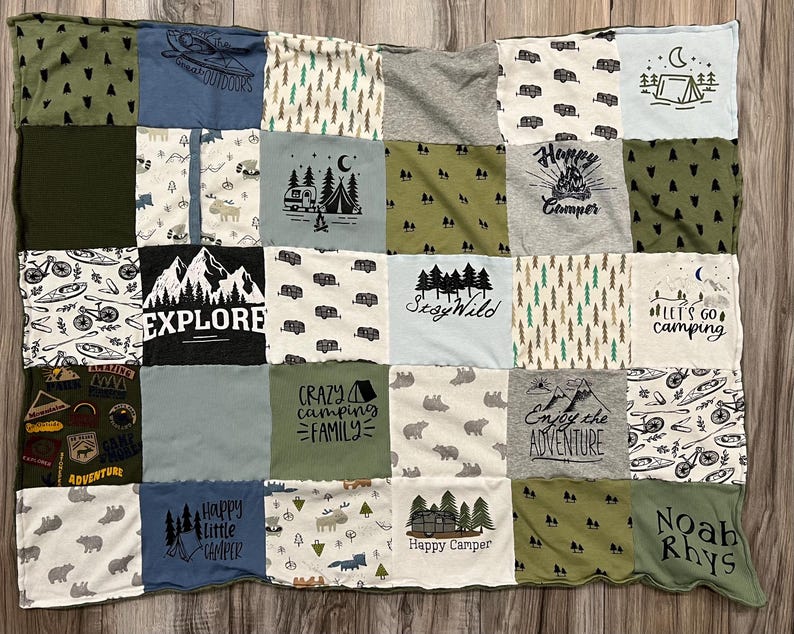 May include: A patchwork quilt made from various t-shirts, featuring camping-themed graphics and text. The quilt includes images of tents, trees, bears, and phrases like "Explore" and "Happy Camper". The color palette includes greens, blues, and grays.