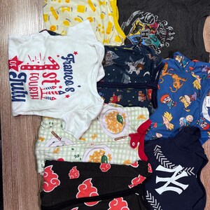May include: A collection of children's clothing items, including t-shirts and onesies. The shirts feature various designs, such as pasta, a Fourth of July graphic, a Harry Potter crest, and a New York Yankees logo. The color palette includes white, blue, gray, and black.