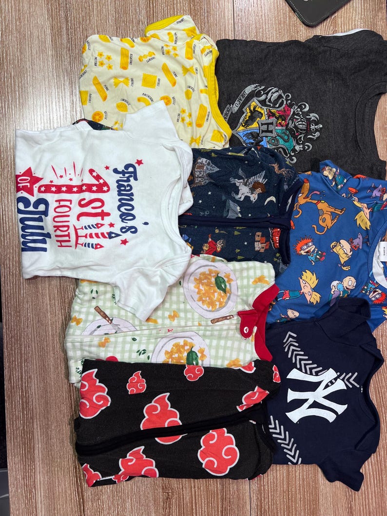 May include: A collection of children's clothing items, including t-shirts and onesies. The shirts feature various designs, such as pasta, a Fourth of July graphic, a Harry Potter crest, and a New York Yankees logo. The color palette includes white, blue, gray, and black.
