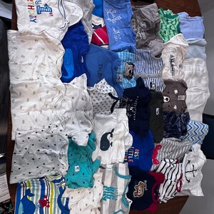 May include: A collection of baby onesies and shirts in various colors and patterns, including stripes, whales, and anchors. Some items have text that reads "My 1st Birthday" and "Best Little". The clothing is arranged on a wooden surface.