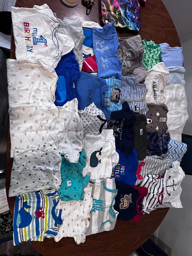 May include: A collection of baby onesies and shirts in various colors and patterns, including stripes, whales, and anchors. Some items have text that reads "My 1st Birthday" and "Best Little". The clothing is arranged on a wooden surface.