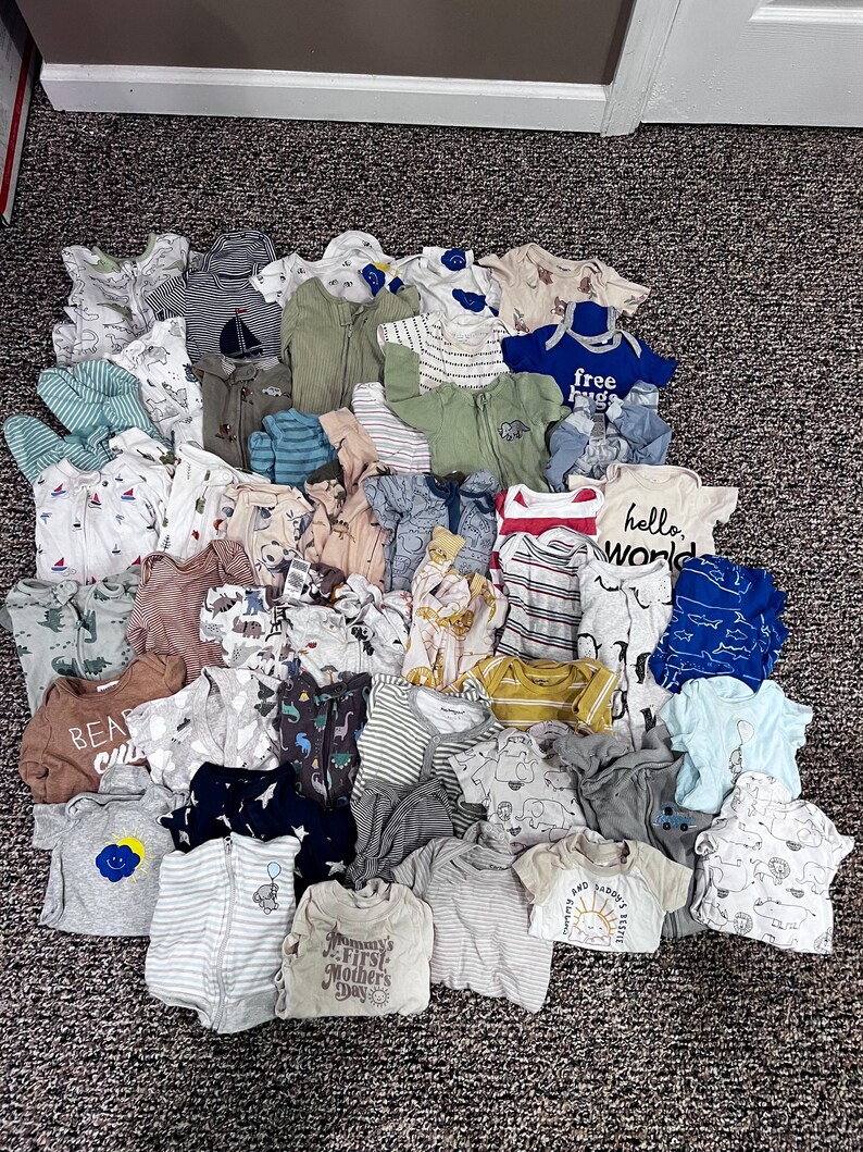 May include: A collection of baby clothes in various colors and styles. The assortment includes onesies, shirts, and zip-up sleepers. Some items feature prints like dinosaurs, sailboats, and the text "hello world".
