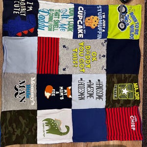 May include: A patchwork quilt made from various children's t-shirts. The shirts feature colorful graphics and text, including phrases like "I'm Roarly Cute," "Born to be Wild," and "Mommy's Little Man." The quilt is a mix of colors and patterns.