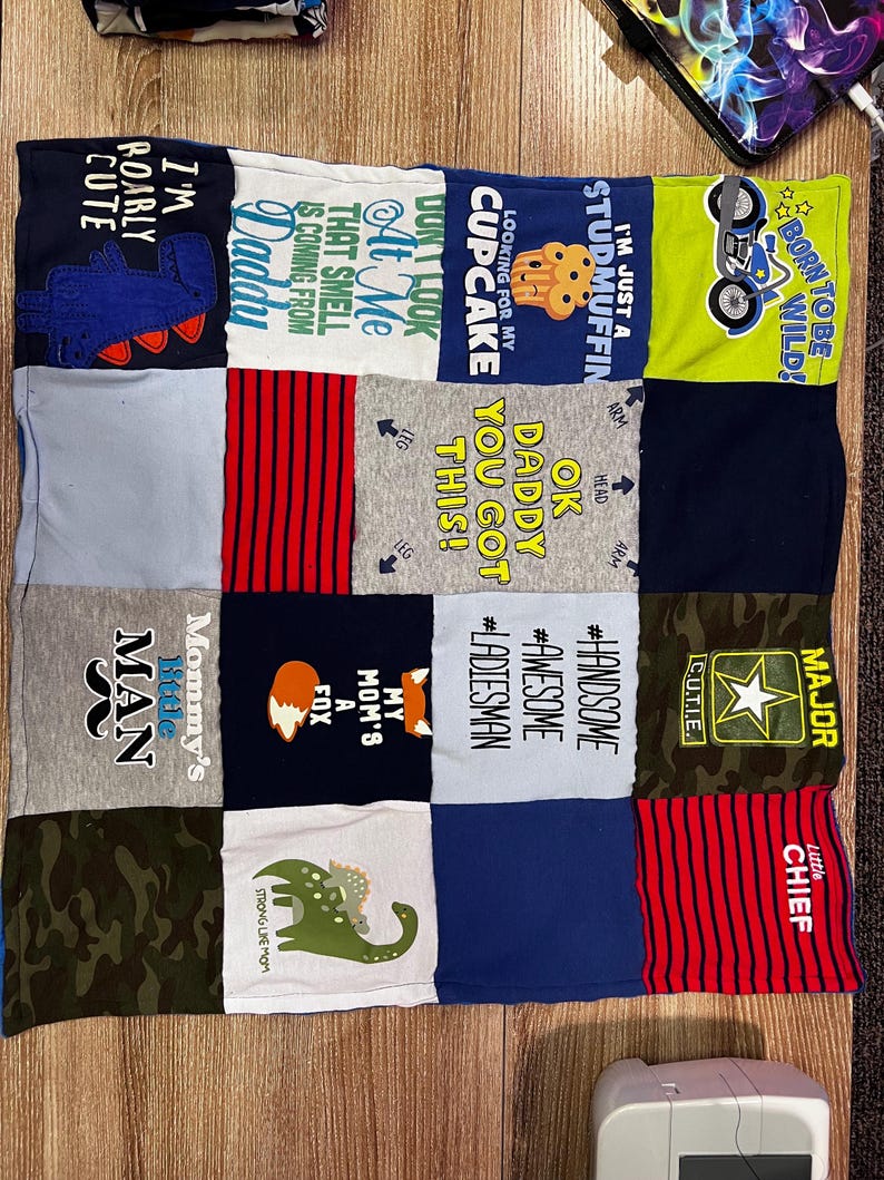 May include: A patchwork quilt made from various children's t-shirts. The shirts feature colorful graphics and text, including phrases like "I'm Roarly Cute," "Born to be Wild," and "Mommy's Little Man." The quilt is a mix of colors and patterns.