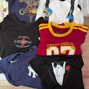 May include: A collection of baby t-shirts and onesies in various colors. Designs include a race car, a NASA patch, a tuxedo, and a "Gryffindor" shirt. Other shirts feature the text "Barrett-Jackson" and "The American Cigarette Offshore Legend".