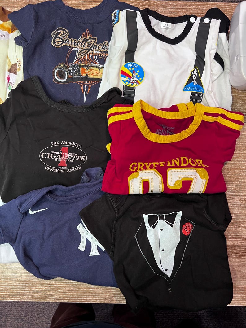 May include: A collection of baby t-shirts and onesies in various colors. Designs include a race car, a NASA patch, a tuxedo, and a "Gryffindor" shirt. Other shirts feature the text "Barrett-Jackson" and "The American Cigarette Offshore Legend".