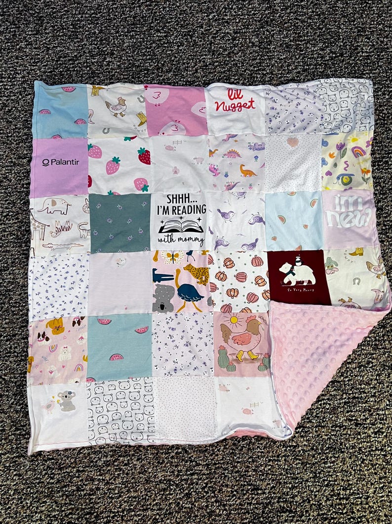 May include: A patchwork baby blanket with various fabric squares in pastel colors. The squares feature prints of animals, strawberries, and the text "lil Nugget" and "SHHH... I'M READING with mommy". The blanket has a pink, textured backing.