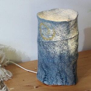 May include: A cylindrical lamp with a blue felt shade and a yellow design. The lamp has a warm glow emanating from within. The base is a copper ring, and the lamp sits on a wooden surface. The lamp is plugged in with a white cord.