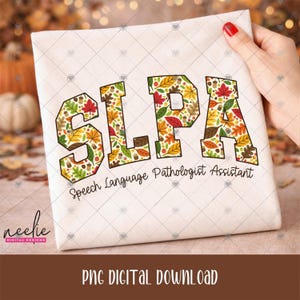May include: A white t-shirt featuring the text SLPA in large block letters filled with a colorful autumn pattern of leaves and acorns. Below the letters, the text Speech Language Pathologist Assistant is printed in a black cursive font.