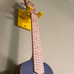 Może przedstawiać: Żółty uchwyt ścienny na ukulele z wiszącym na nim ukulele. Ukulele ma ciemnoniebieskie ciało i drewniany gryf.