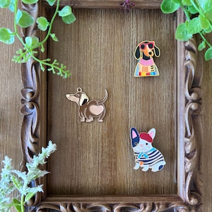 May include: Three enamel pins featuring dog designs. One pin shows a dachshund wearing red sunglasses and a colorful sweater. Another pin depicts a brown dachshund with a heart-shaped patch on its rear. The third pin is a French bulldog wearing a red beret and a blue and white striped shirt.