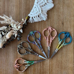 May include: Five decorative embroidery scissors in various colors, including iridescent, rose gold, and blue, are arranged on a wooden surface. The scissors have ornate handles and sharp, silver blades. A macrame piece and dried flowers are in the background.